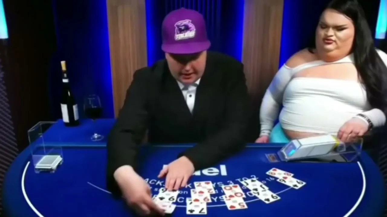 A Duel blackjack dealer in a suit and a purple baseball cap with a guest beside him